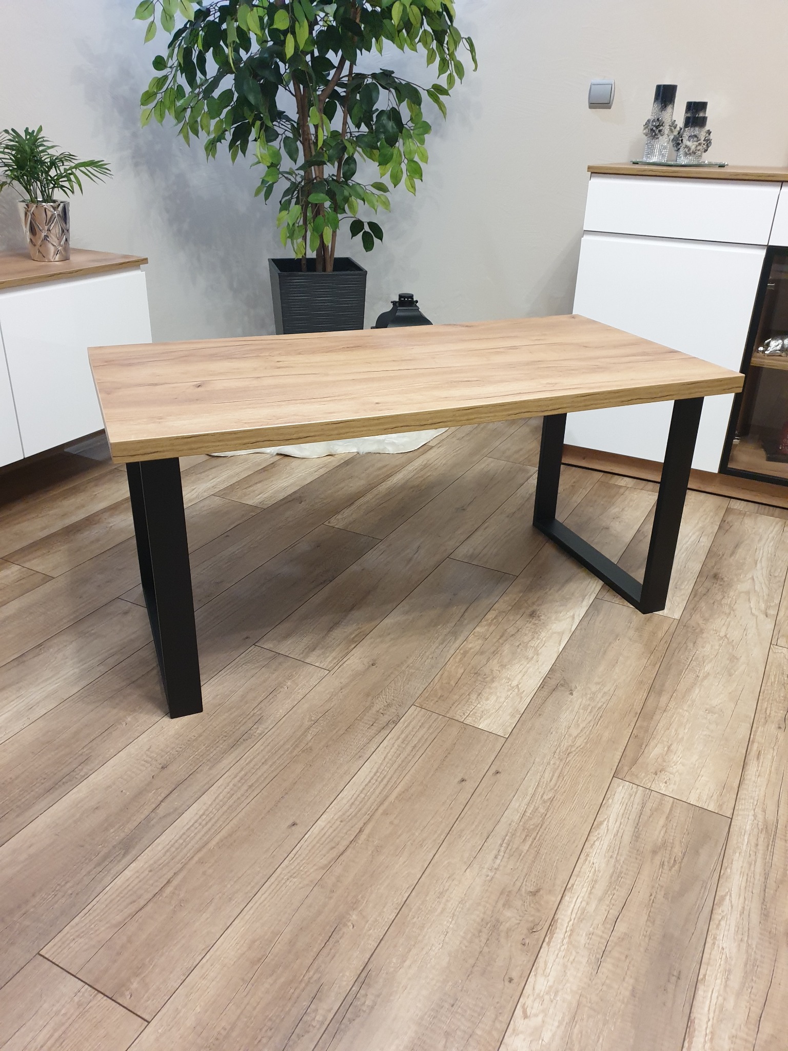 Stół z blatem imitującym jasne drewno i czarnymi, metalowymi nogami w kształcie litery U, na drewnianej podłodze, w tle rośliny doniczkowe i białe szafki.