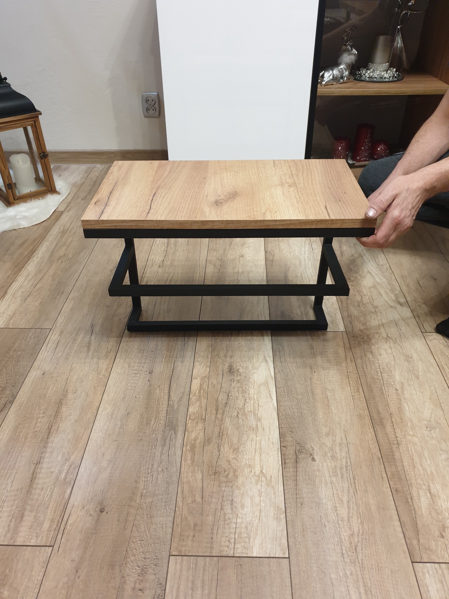 Stolik kawowy z blatem imitującym drewno i czarną, metalową podstawą, umieszczony na podłodze z paneli. Widoczne ręce osoby przesuwającej stolik.