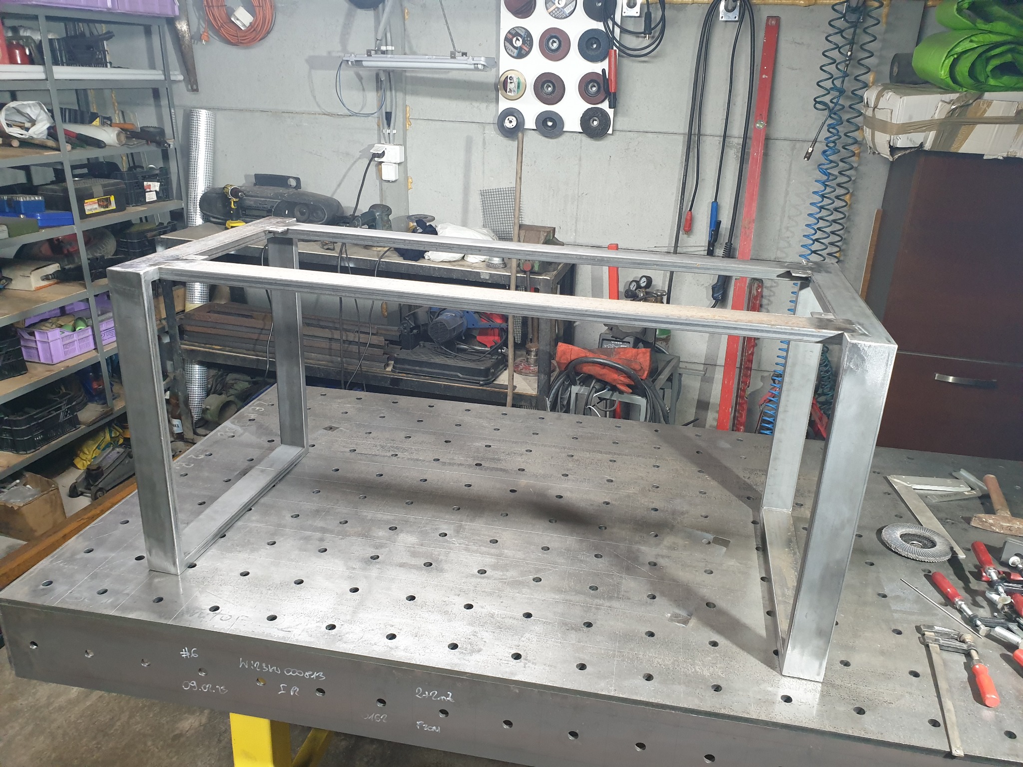 Konstrukcja ramy stołu ze stali, spawana na perforowanym stole spawalniczym w warsztacie, widoczne narzędzia i akcesoria spawalnicze w tle.