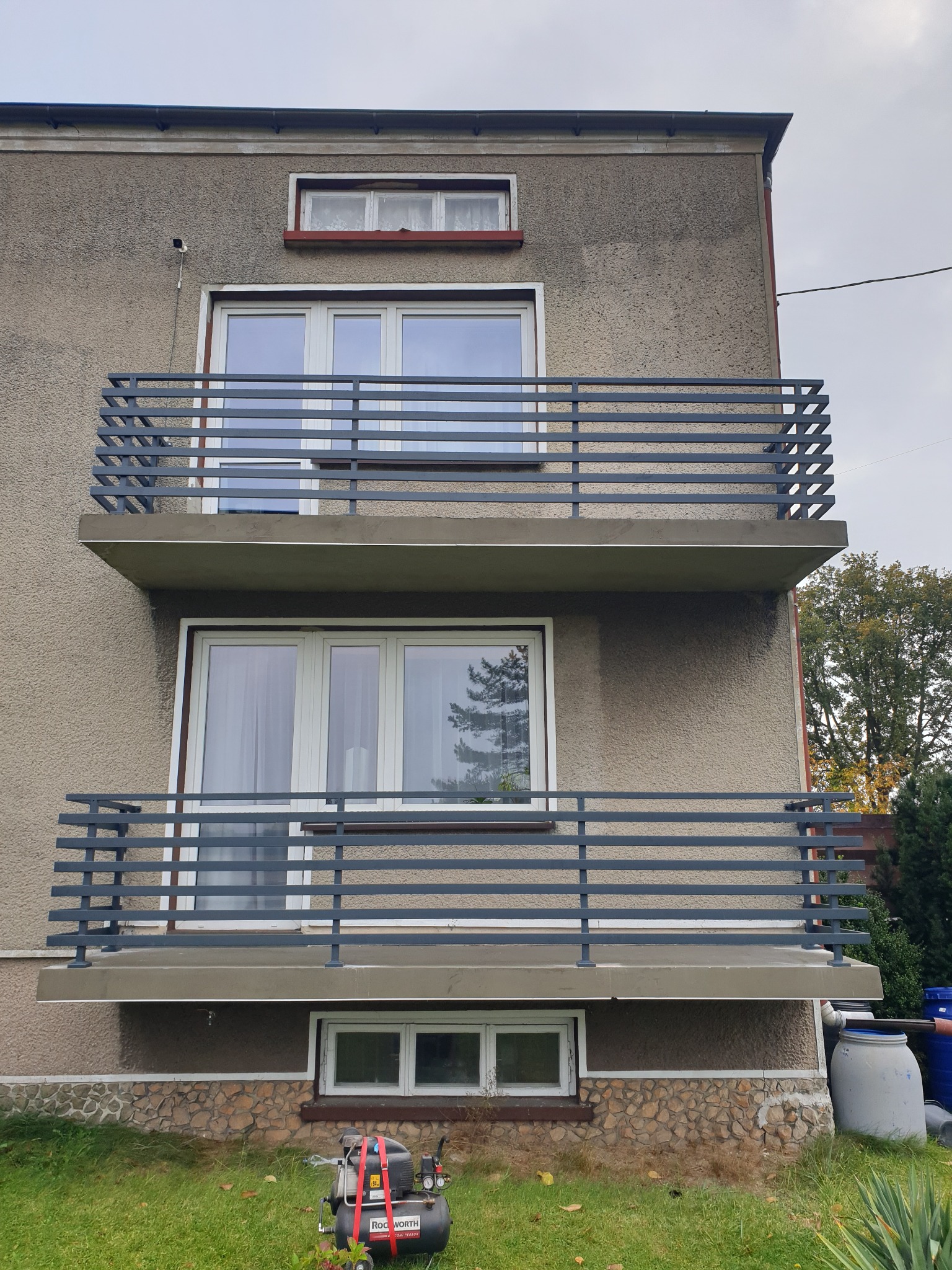 Dwu-kondygnacyjny budynek z balkonami zabezpieczonymi szarą balustradą poziomą, na trawniku stoi sprężarka powietrza ROCKWORTH z czerwonymi pasami.