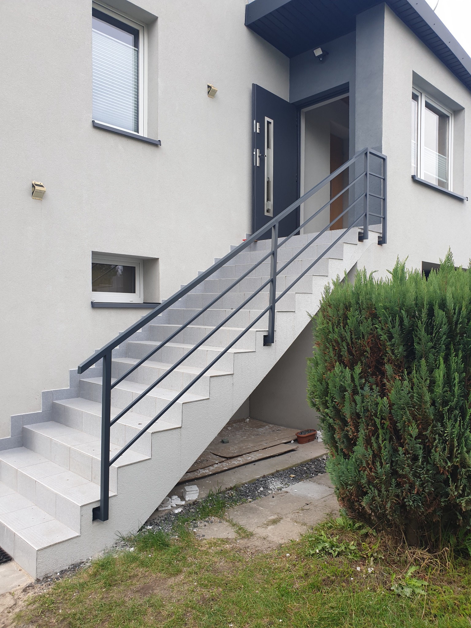 Zewnętrzne schody wejściowe do domu z szarą, minimalistyczną balustradą z poziomych prętów, widok z dołu, krzew iglasty po prawej stronie.