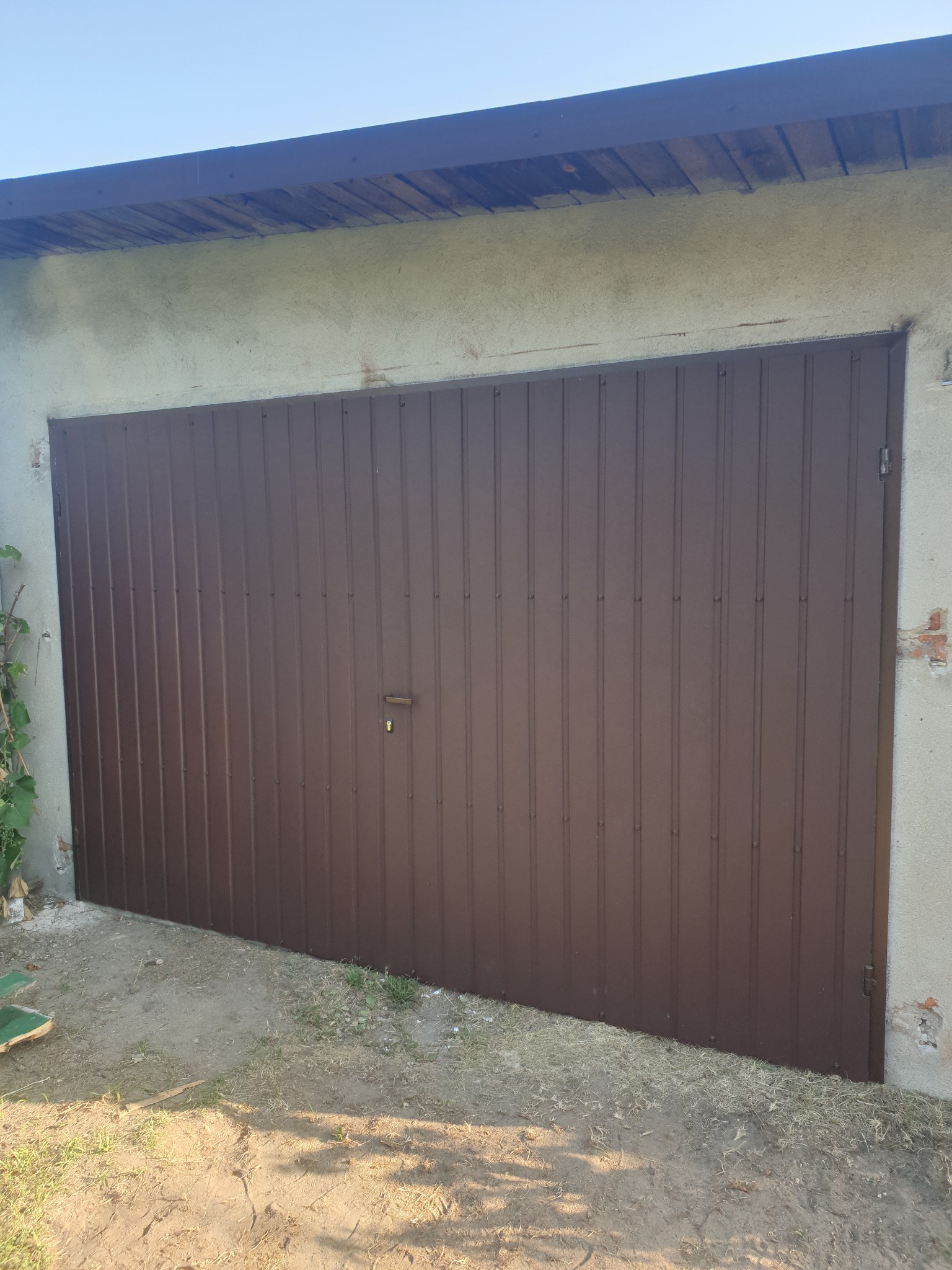 Brązowa brama garażowa z pionowymi przetłoczeniami, zamontowana w szarym, lekko uszkodzonym murze, widziana z perspektywy lekko z dołu.