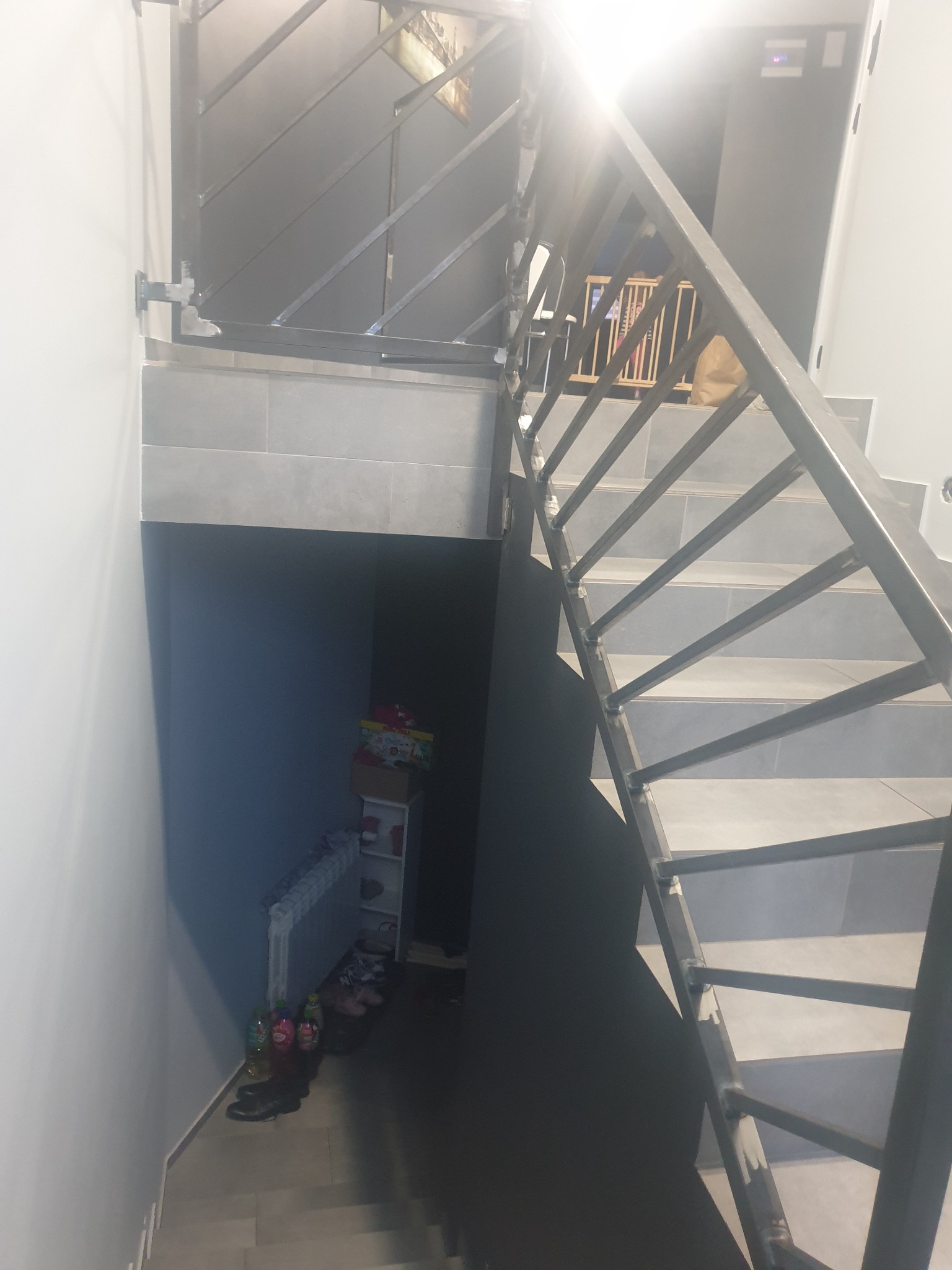 Metalowe schody z surową, spawaną balustradą wewnątrz domu, widok z góry na dół, z widocznym fragmentem piwnicy/schowka pod schodami.