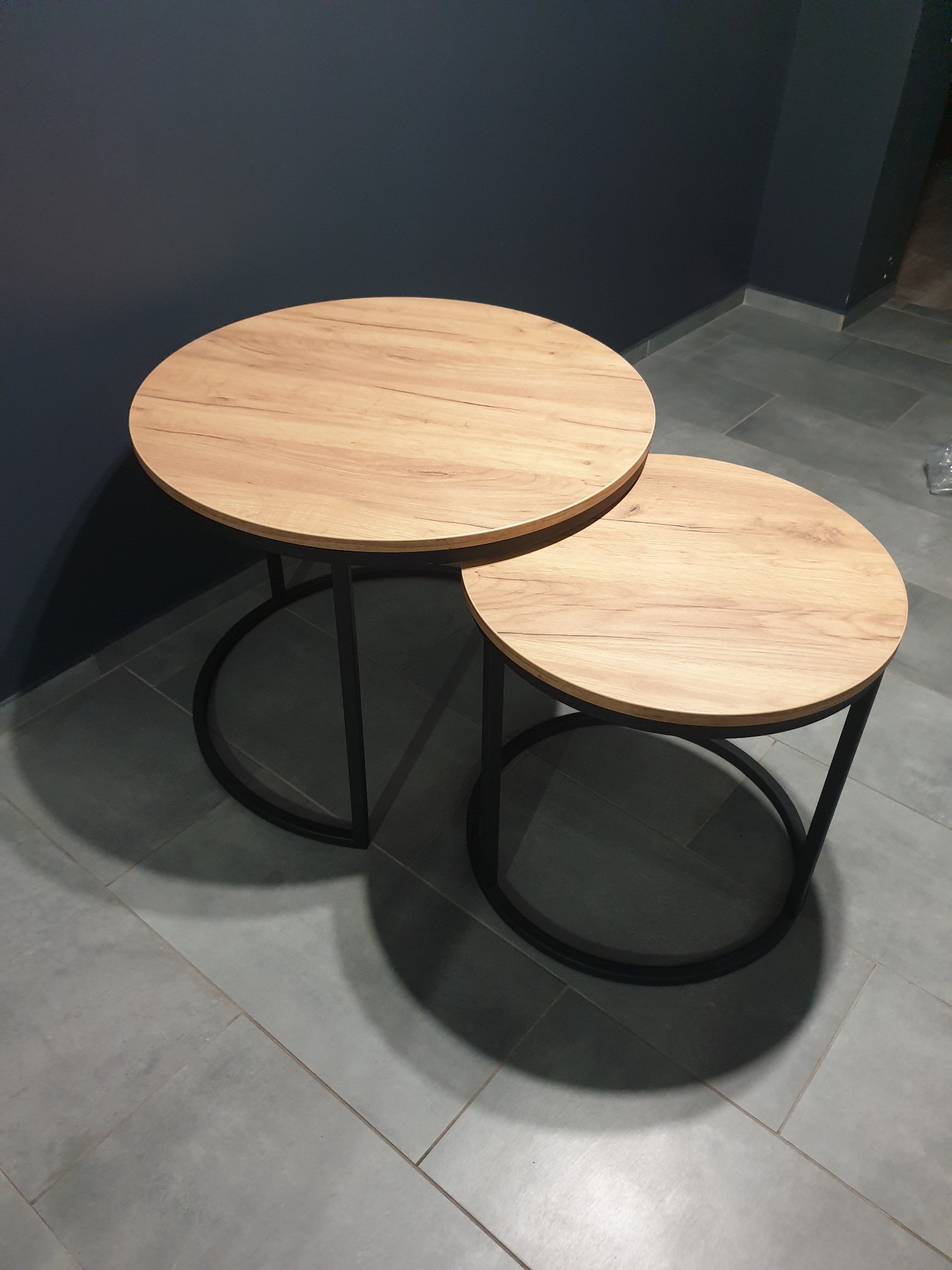 Dwa okrągłe stoliki kawowe z drewnianymi blatami i czarnymi metalowymi nogami, ustawione jeden obok drugiego na szarych płytkach w Blachowni, na tle ciemnej ściany.