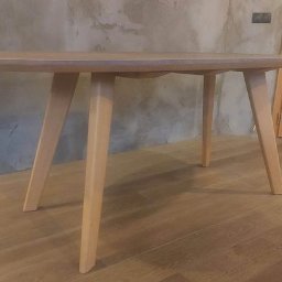 AT Tomaszewski - Owalny stolik kawowy z jasnego drewna na trzech nogach, prezentowany w warszawskim wnętrzu z surową, betonową ścianą i drewnianą podłogą.