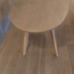 AT Tomaszewski - Owalny stolik kawowy z jasnego drewna na trzech nogach, ustawiony na drewnianej podłodze, widoczne drzwi w tle.