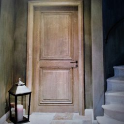 AT Tomaszewski - Drewniane drzwi wewnętrzne w klasycznym stylu, z dekoracyjną ramą i czarną klamką, oświetlone boczną lampą z zapaloną świecą, widoczne fragmenty schodów.