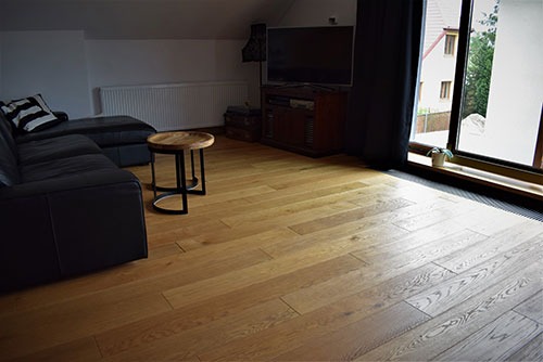 Jasne drewniane deski podłogowe ułożone w jodełkę w pokoju na poddaszu, widoczny narożnik sofy, mały okrągły stolik kawowy i okno balkonowe z rośliną.