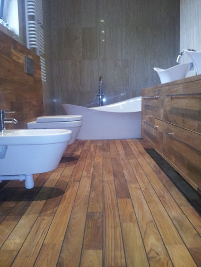 Bliższe ujęcie drewnianej podłogi w łazience z białymi elementami armatury i umywalką na szafce z drewna.