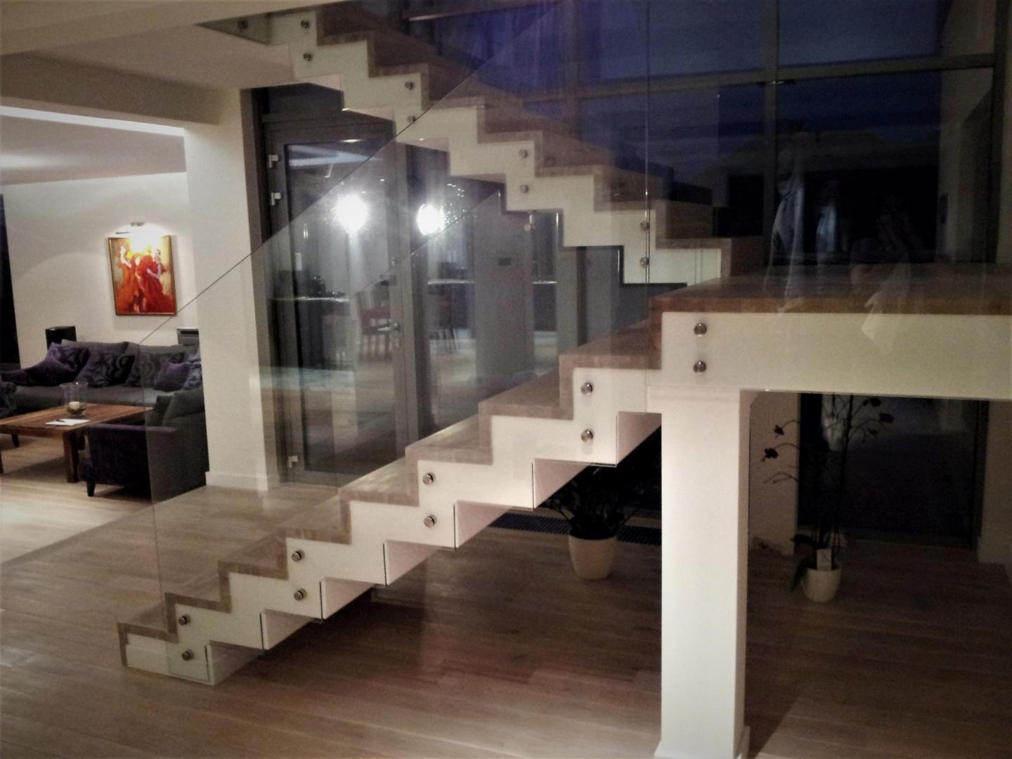 Nowoczesne schody wspornikowe z drewnianymi stopniami i szklaną balustradą, zamocowane na metalowych śrubach, w przestronnym wnętrzu z parkietem i widocznym salonem w tle.