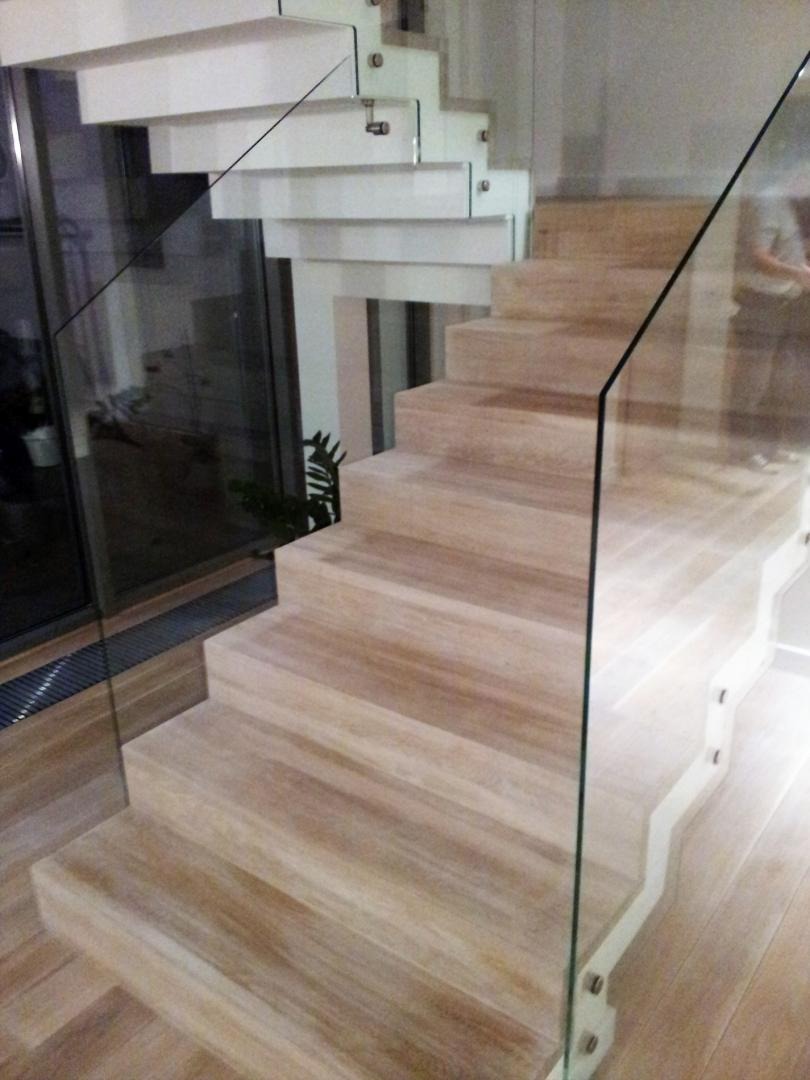 Nowoczesne schody z jasnego drewna z białymi podstopnicami i szklaną balustradą, widok od dołu, z widocznym fragmentem okna w tle.