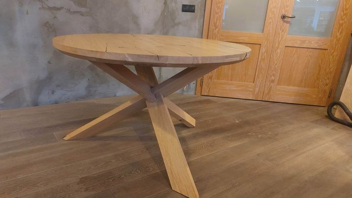 Okrągły stół z jasnego drewna z charakterystycznymi pęknięciami na blacie, osadzony na geometrycznej podstawie z krzyżujących się nóg, na tle surowej ściany i drewnianych drzwi.