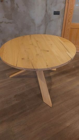 Okrągły stół z jasnego drewna z widocznymi naturalnymi pęknięciami i sękami, wsparty na trzech ukośnych nogach, na tle betonowej ściany i fragmentu drewnianych drzwi.