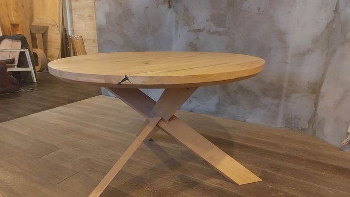 Okrągły stół z jasnego drewna z charakterystycznym pęknięciem na blacie, wsparty na krzyżujących się nogach, na tle drewnianej podłogi i surowej ściany.