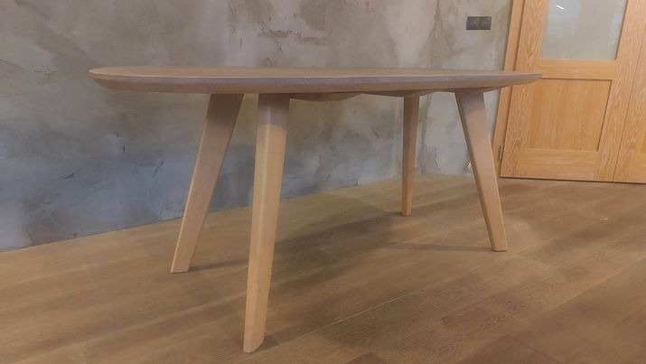 Owalny stolik kawowy z jasnego drewna na trzech nogach, prezentowany w warszawskim wnętrzu z surową, betonową ścianą i drewnianą podłogą.