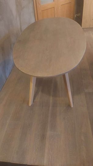 Owalny stolik kawowy z jasnego drewna na trzech nogach, ustawiony na drewnianej podłodze, widoczne drzwi w tle.