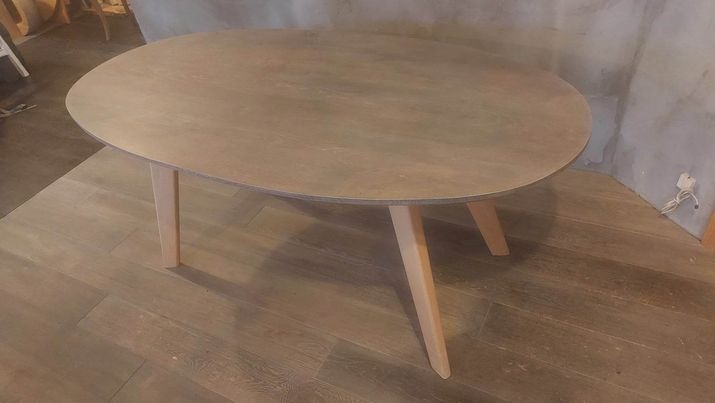 Owalny stolik kawowy z blatem w odcieniach szarości i jasnymi, drewnianymi nogami, ustawiony na podłodze z paneli w ciepłym odcieniu brązu, na tle surowej, betonowej ściany.