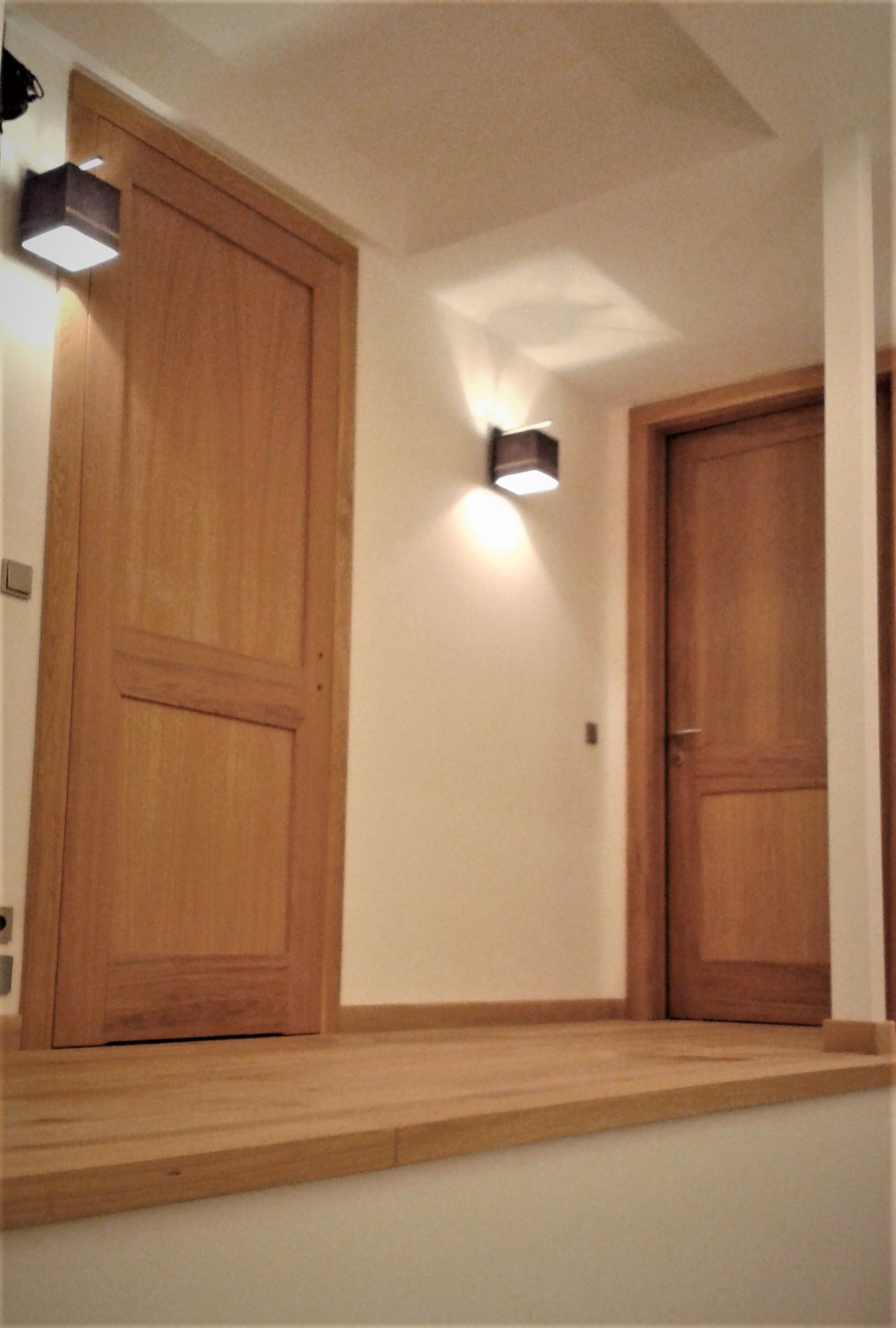 Dwa drewniane drzwi wewnętrzne z ościeżnicami, oświetlone kwadratowymi kinkietami, zamontowane na ścianie w korytarzu z podwyższoną drewnianą podłogą.