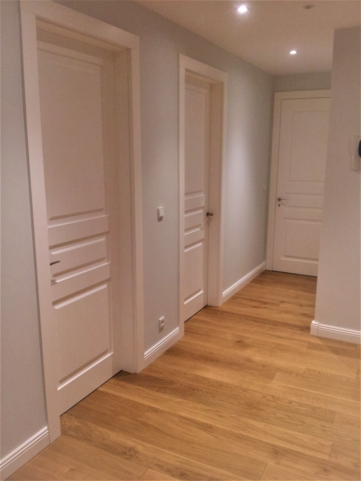 Korytarz z trzema białymi, wewnętrznymi drzwiami z poziomymi frezowaniami i podłogą z paneli w odcieniach brązu, oświetlony punktowymi lampami sufitowymi.