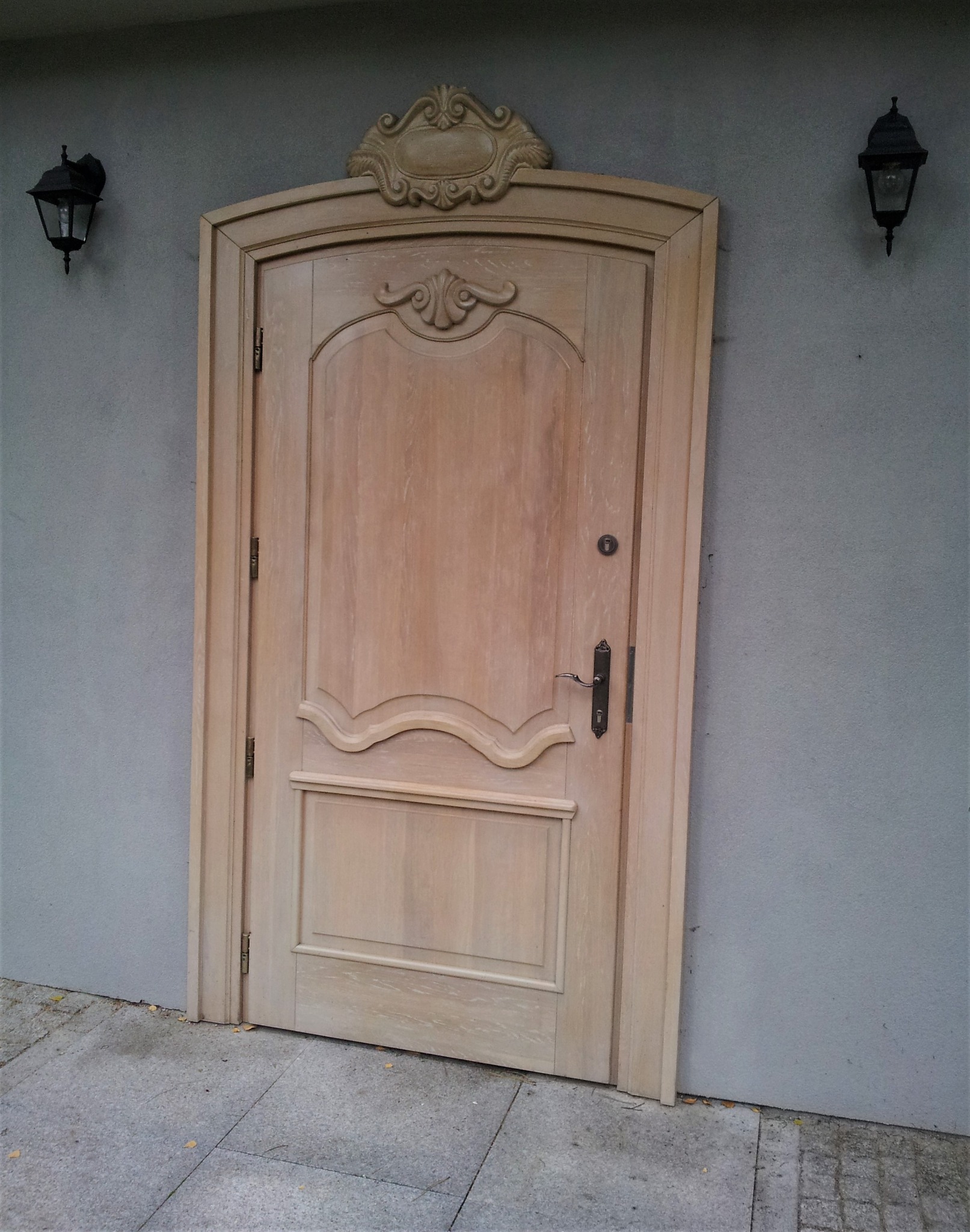 Jasna, drewniana brama wejściowa z ozdobnymi elementami, zamontowana w szarej ścianie z dwoma czarnymi lampami po bokach.