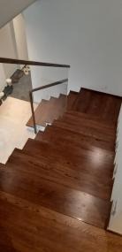 Wysoki kąt widzenia na ciemnobrązowe schody z drewnianymi stopniami i metalową balustradą, prowadzące w dół do jasnego pomieszczenia.