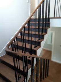 Nowoczesne schody z drewnianymi stopniami i balustradą z czarnymi, pionowymi szczebelkami oraz jasnobrązową poręczą, widok z dołu.