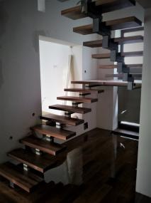 Nowoczesne schody z ciemnego drewna z metalowymi wspornikami, zawieszone w przestrzeni, widoczne elementy montażowe, surowy stan wykończenia wnętrza.