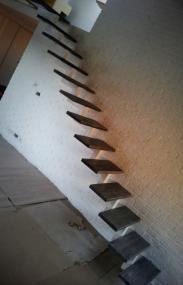 Stopnie schodów wspornikowych z ciemnego drewna, zamocowane do białej, teksturowanej ściany. Widoczne surowe wykończenie i elementy montażowe, sugerujące etap budowy.