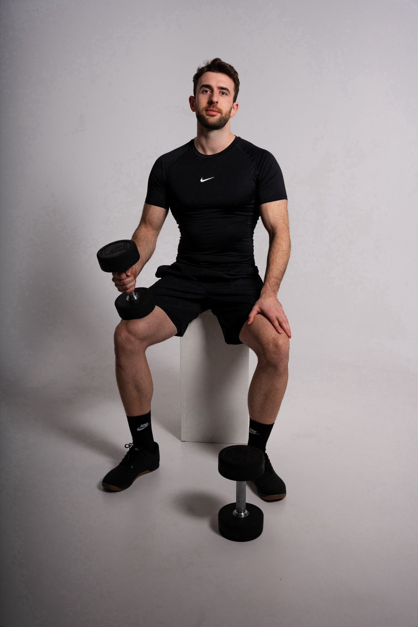 Mężczyzna w czarnym stroju sportowym Nike siedzi na białym sześcianie, trzymając hantlę w prawej ręce, druga hantla stoi na podłodze obok jego nogi.
