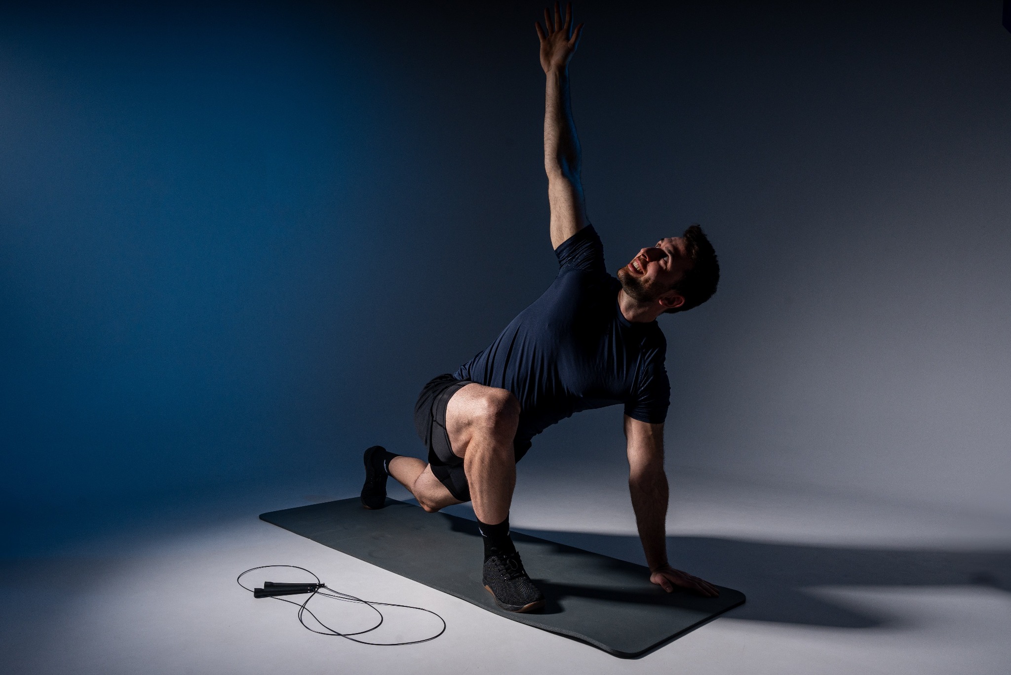 Mężczyzna w czarnym stroju sportowym wykonuje dynamiczne rozciąganie na szarej macie, unosząc jedną rękę ku górze; w tle dwukolorowe oświetlenie i skakanka.
