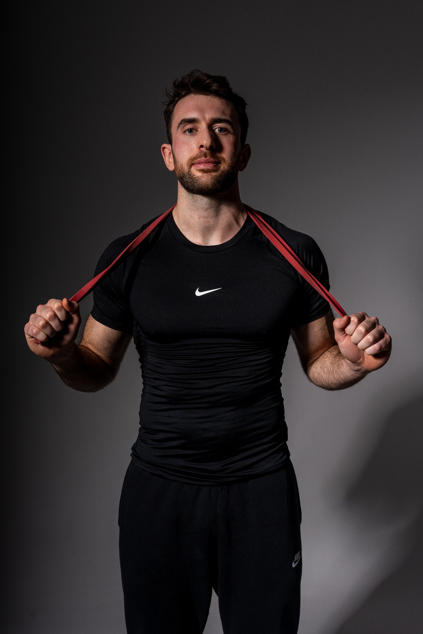 Mężczyzna w czarnym sportowym stroju trzyma czerwoną gumę do ćwiczeń na tle szarego studia.