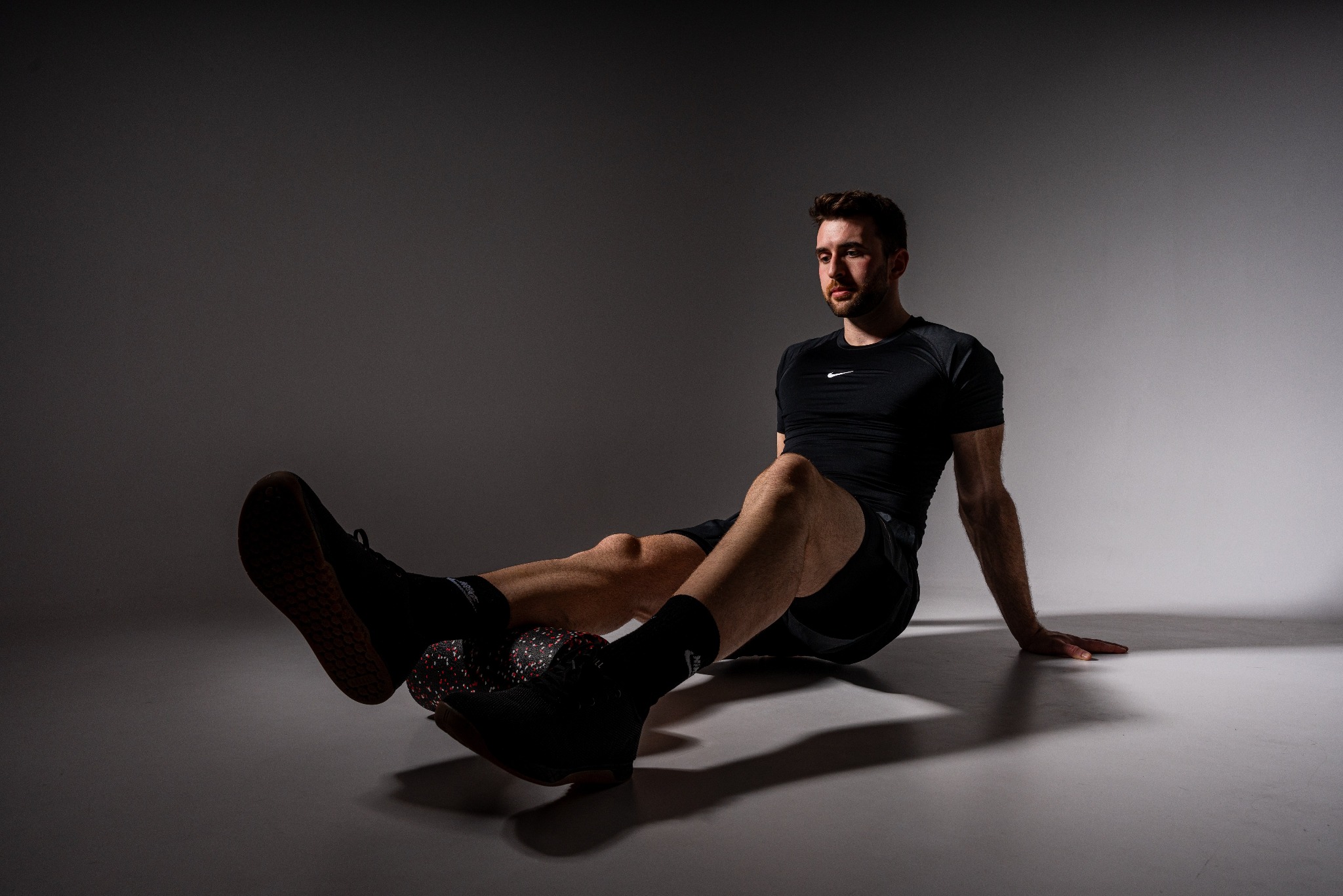 Mężczyzna w sportowym stroju, siedząc na podłodze, wykonuje ćwiczenie z rollerem do masażu nóg, patrząc w bok, z dłonią opartą o podłoże.