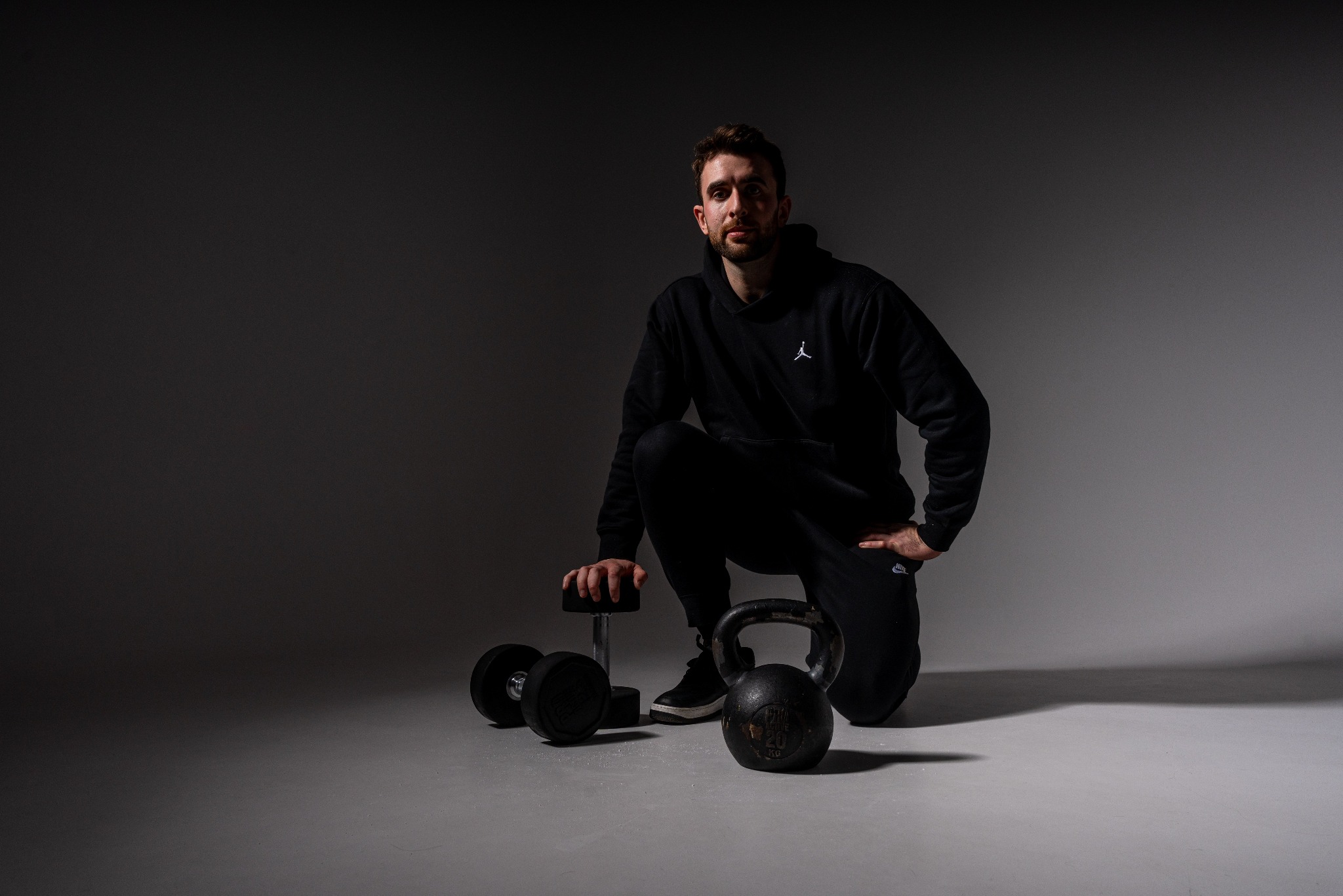 Młody mężczyzna w czarnym dresie, w przysiadzie obok kettlebell 20 kg i hantli na tle szarego studia.