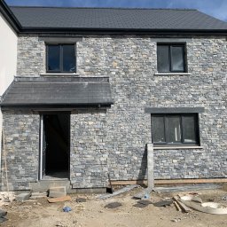 VIKING - Dwukondygnacyjny dom w trakcie obkładania elewacji szarą kamienną okładziną, widoczne okna z czarnymi ramami, szare dachówki, oraz bałagan budowlany wokół budynku.