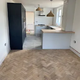 Jasny parkiet ułożony w jodełkę w nowo wyremontowanej kuchni z szarymi szafkami i drewnianym blatem, widoczne wiszące lampy z abażurami ze sznurka i czarna szafa.