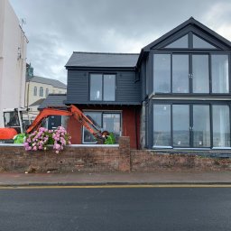 VIKING - Dom w trakcie rozbudowy z użyciem małej koparki, widoczna murowana podmurówka i nowoczesna dobudówka z dużymi oknami.