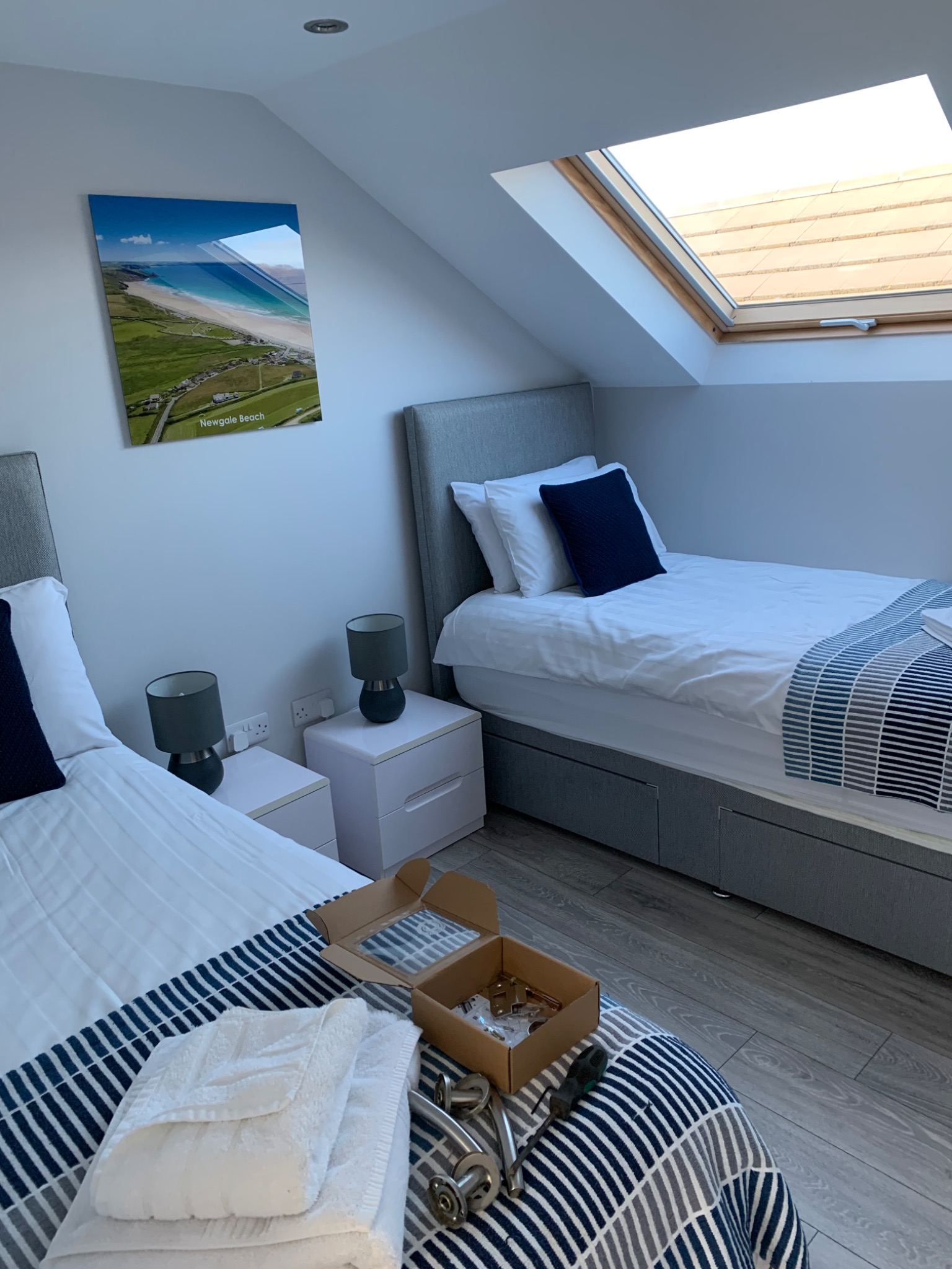 Pokój z dwoma łóżkami, oknem dachowym, lampkami nocnymi i obrazem plaży na ścianie, z rozpakowanymi elementami montażowymi na jednym z łóżek.