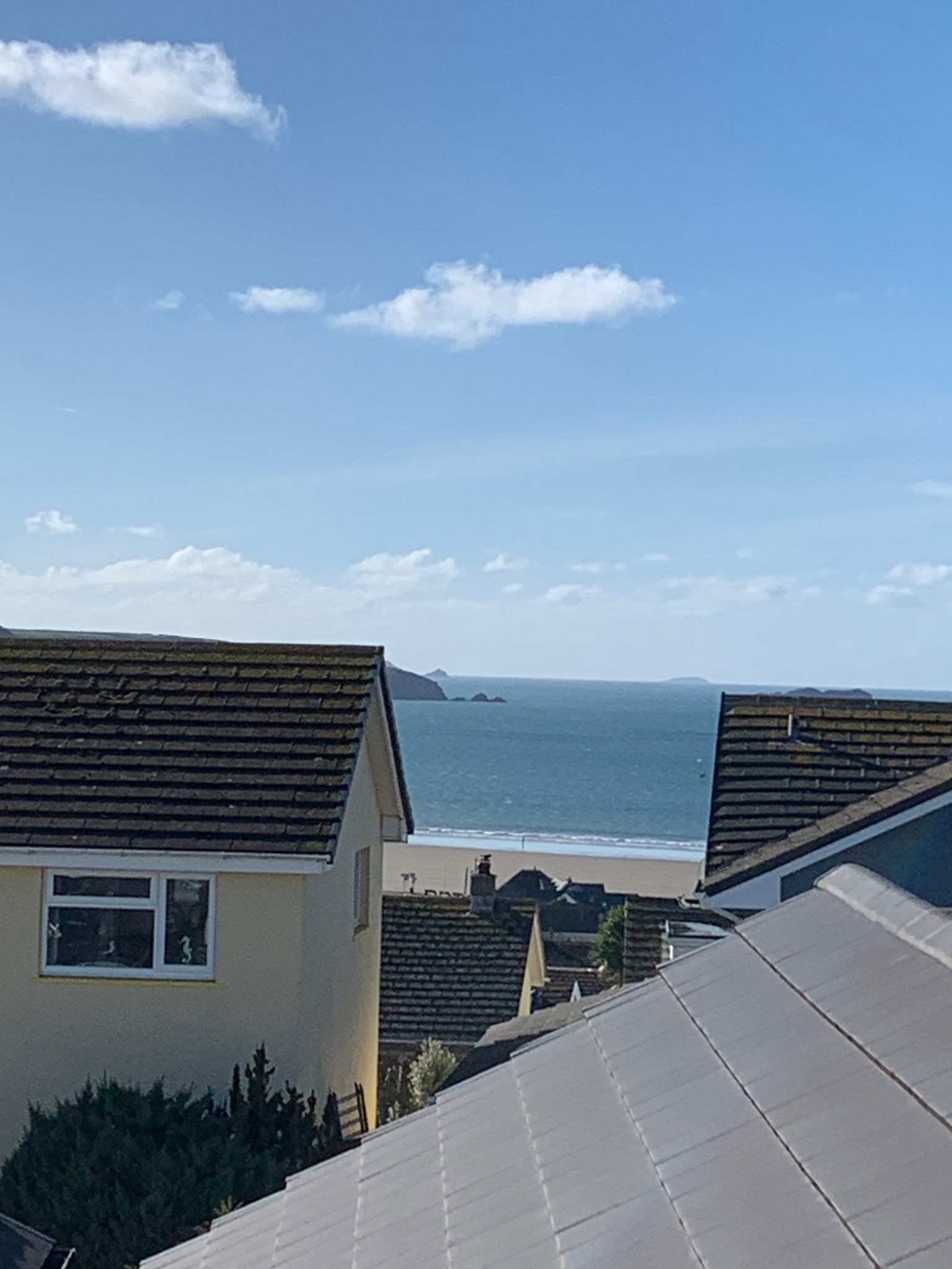 Widok na morze zza dachów budynków w słoneczny dzień, z widocznymi dachówkami i oknami domów.