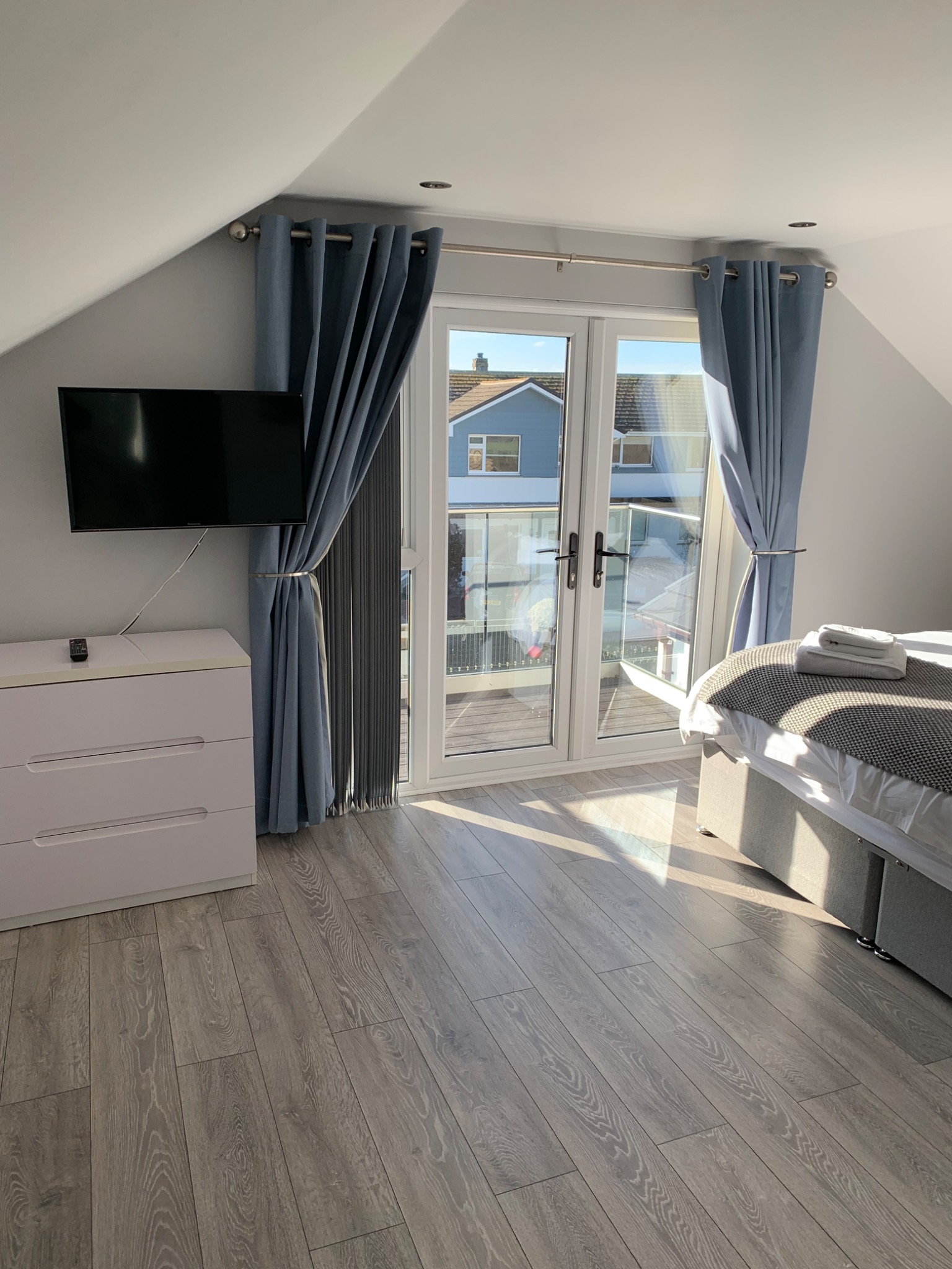 Pokój na poddaszu z szarymi panelami podłogowymi, białą komodą, telewizorem na ścianie, łóżkiem oraz balkonem z widokiem na dom.