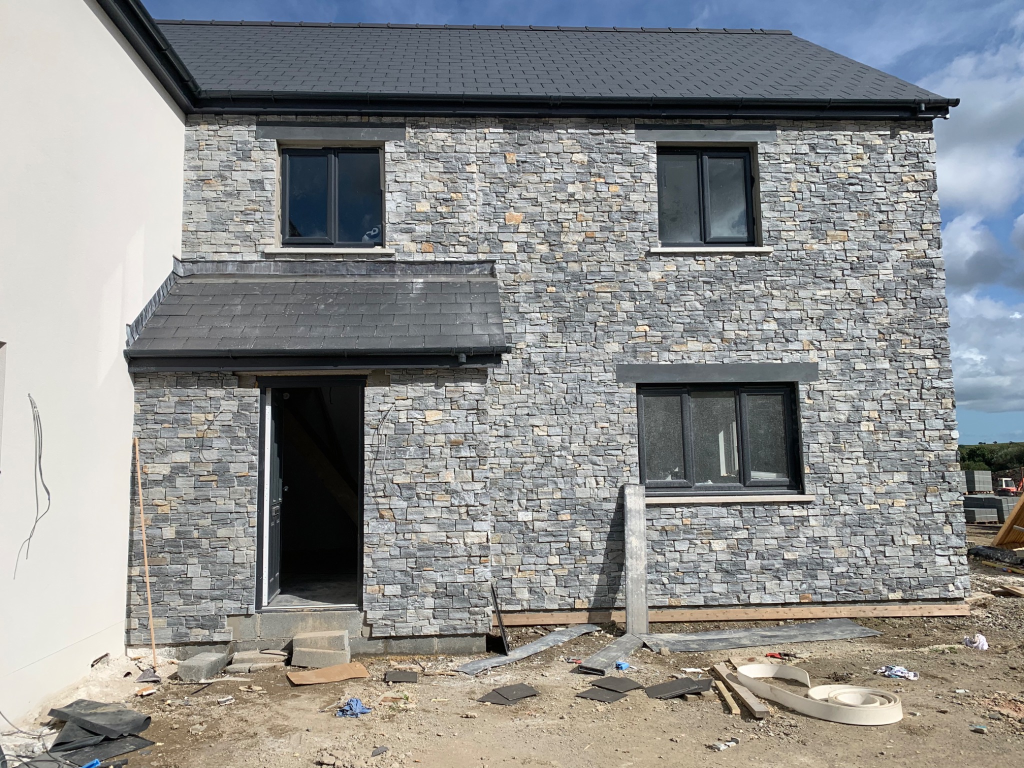 Dwukondygnacyjny dom w trakcie obkładania elewacji szarą kamienną okładziną, widoczne okna z czarnymi ramami, szare dachówki, oraz bałagan budowlany wokół budynku.