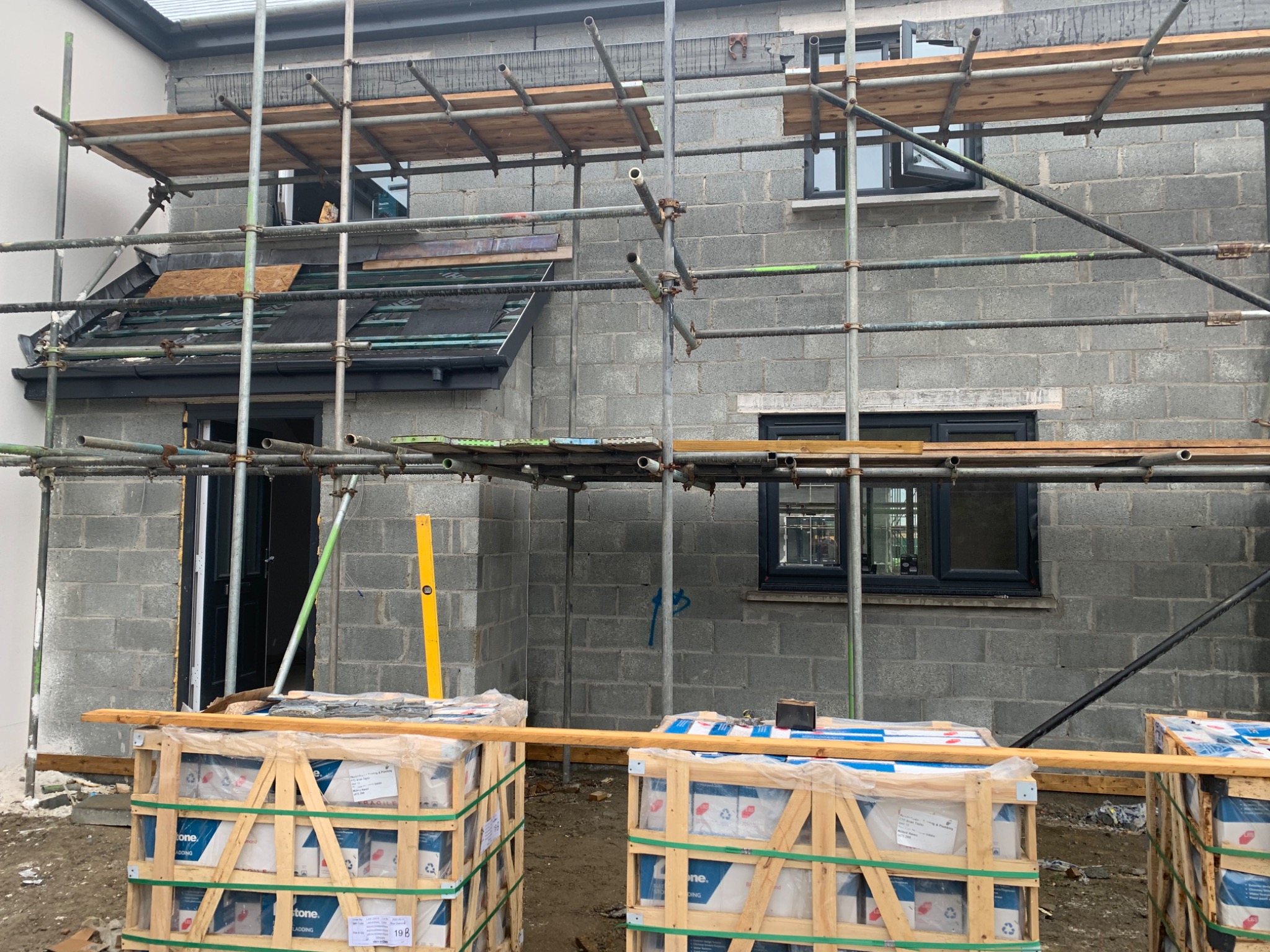 Budynek w stanie surowym zamkniętym z szarych bloczków, widoczne rusztowania, okna, częściowo położone pokrycie dachu i palety z materiałami budowlanymi na pierwszym planie.