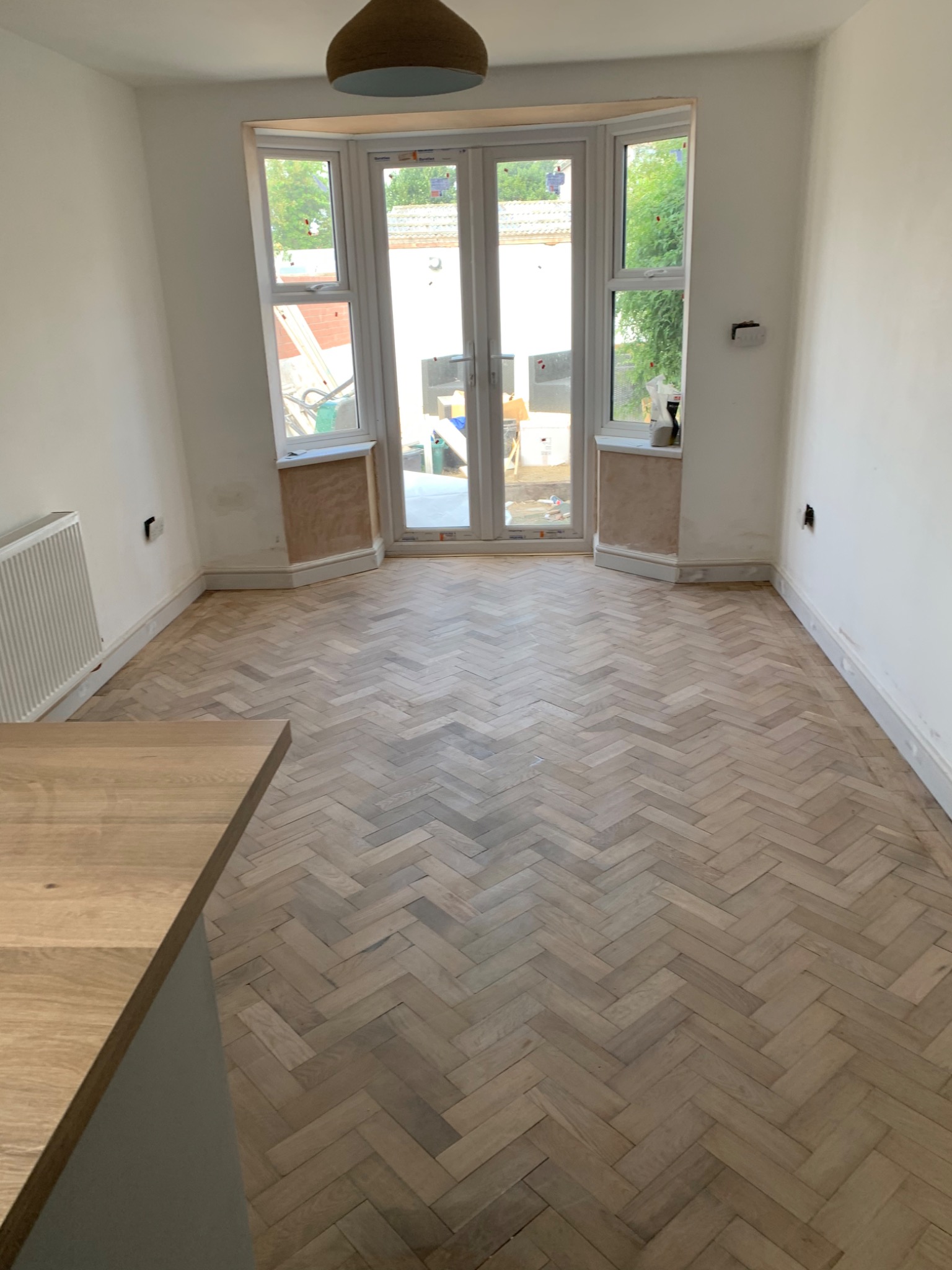 Jasny parkiet ułożony we wzór jodełki w pomieszczeniu w trakcie remontu, widoczne niedokończone obróbki okien i ściany z wystającymi przewodami.