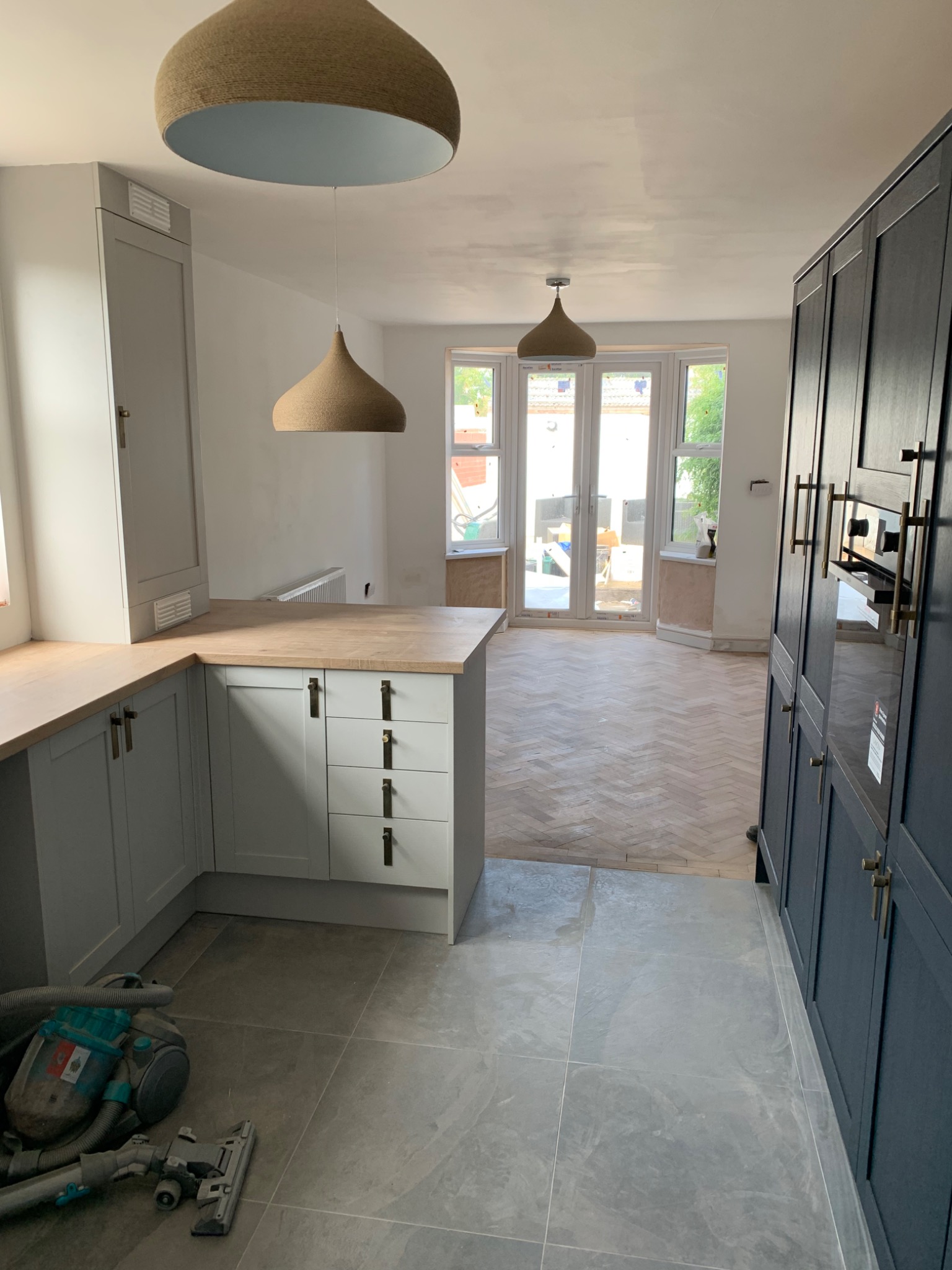 Wnętrze kuchni w trakcie remontu: szare płytki podłogowe, zabudowa kuchenna w odcieniach szarości i granatu z drewnianymi blatami, widoczne ślady szpachlowania na suficie, parkiet w jodełkę...