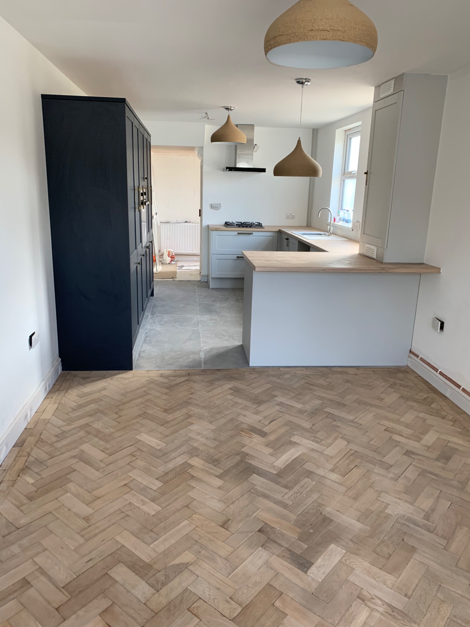 Jasny parkiet ułożony w jodełkę w nowo wyremontowanej kuchni z szarymi szafkami i drewnianym blatem, widoczne wiszące lampy z abażurami ze sznurka i czarna szafa.