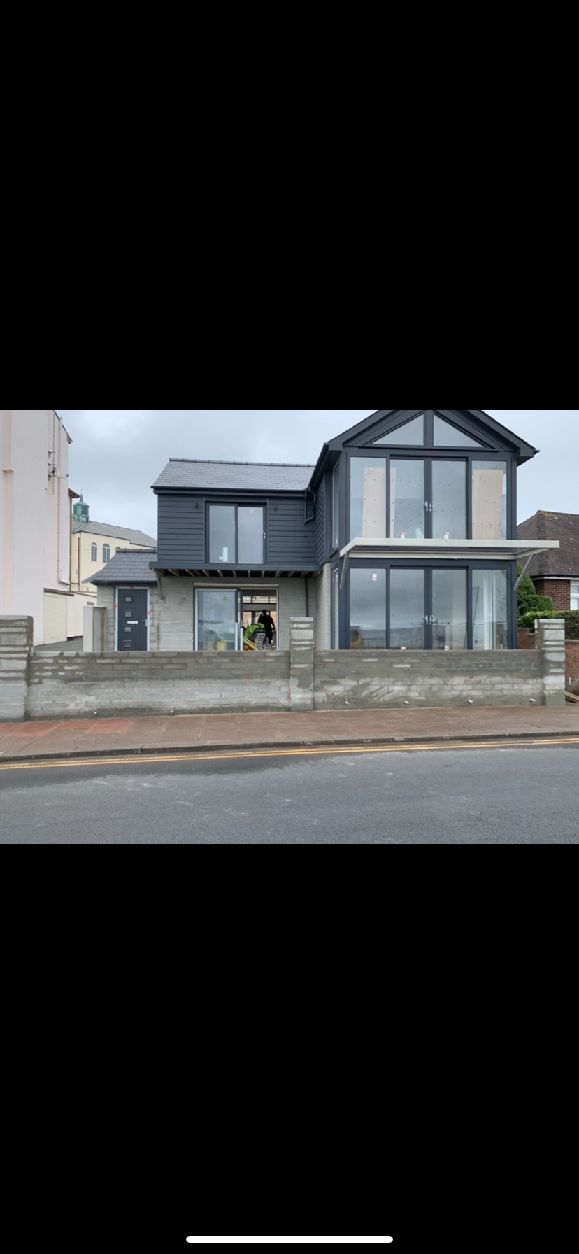 Dwupiętrowy dom z szarą elewacją i dużymi oknami na parterze, w trakcie prac wykończeniowych, widoczny pracownik wewnątrz budynku.