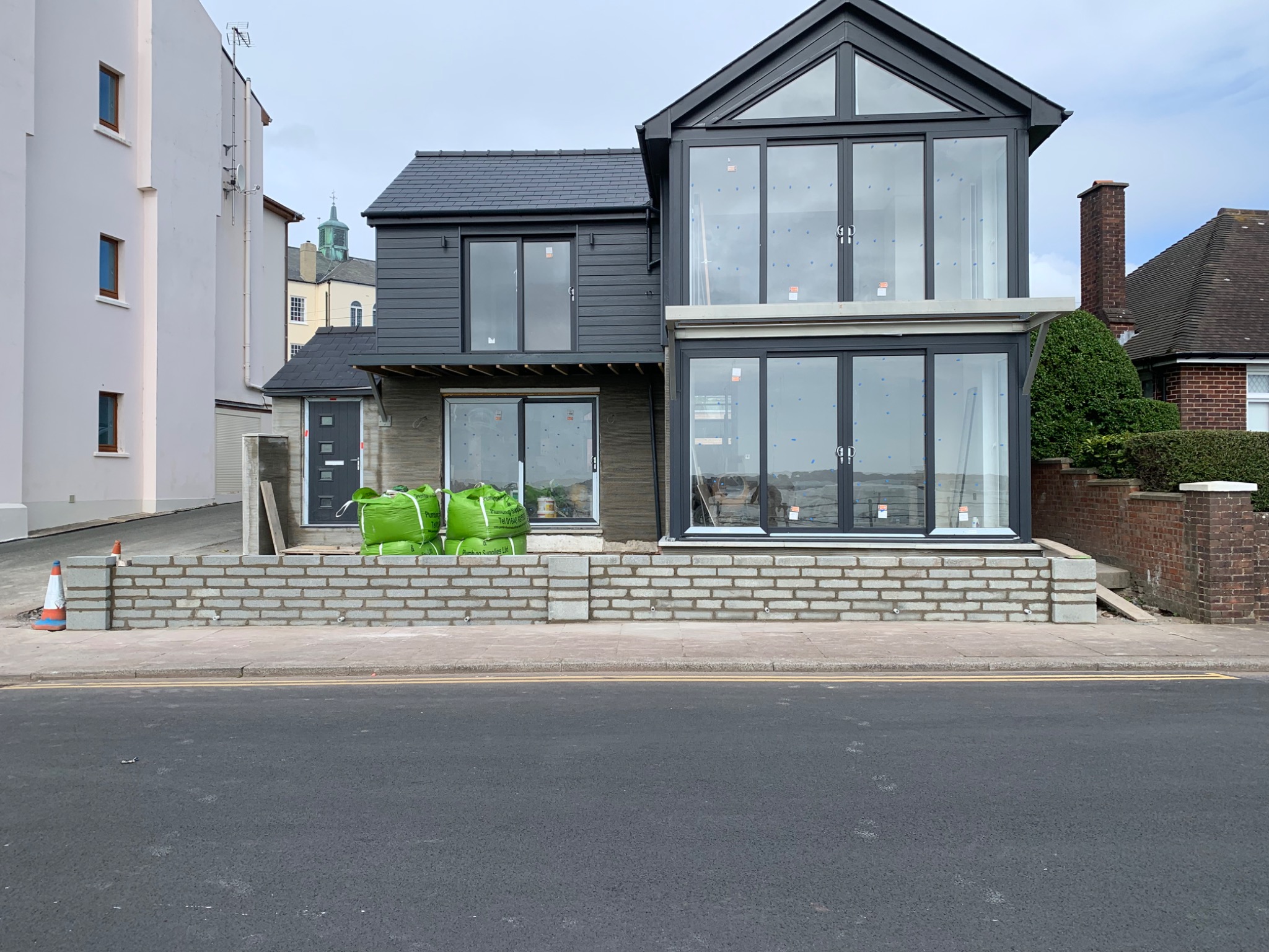 Dom w trakcie budowy z nowoczesną szarą elewacją, dużymi oknami i workami z materiałami budowlanymi przed wejściem.