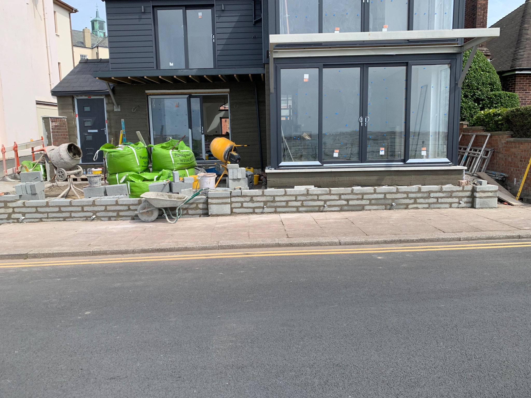 Budowa domu w trakcie, widoczne worki z materiałami budowlanymi, betoniarka, bloczki betonowe i wózek budowlany na tle czarnej elewacji z dużymi oknami.