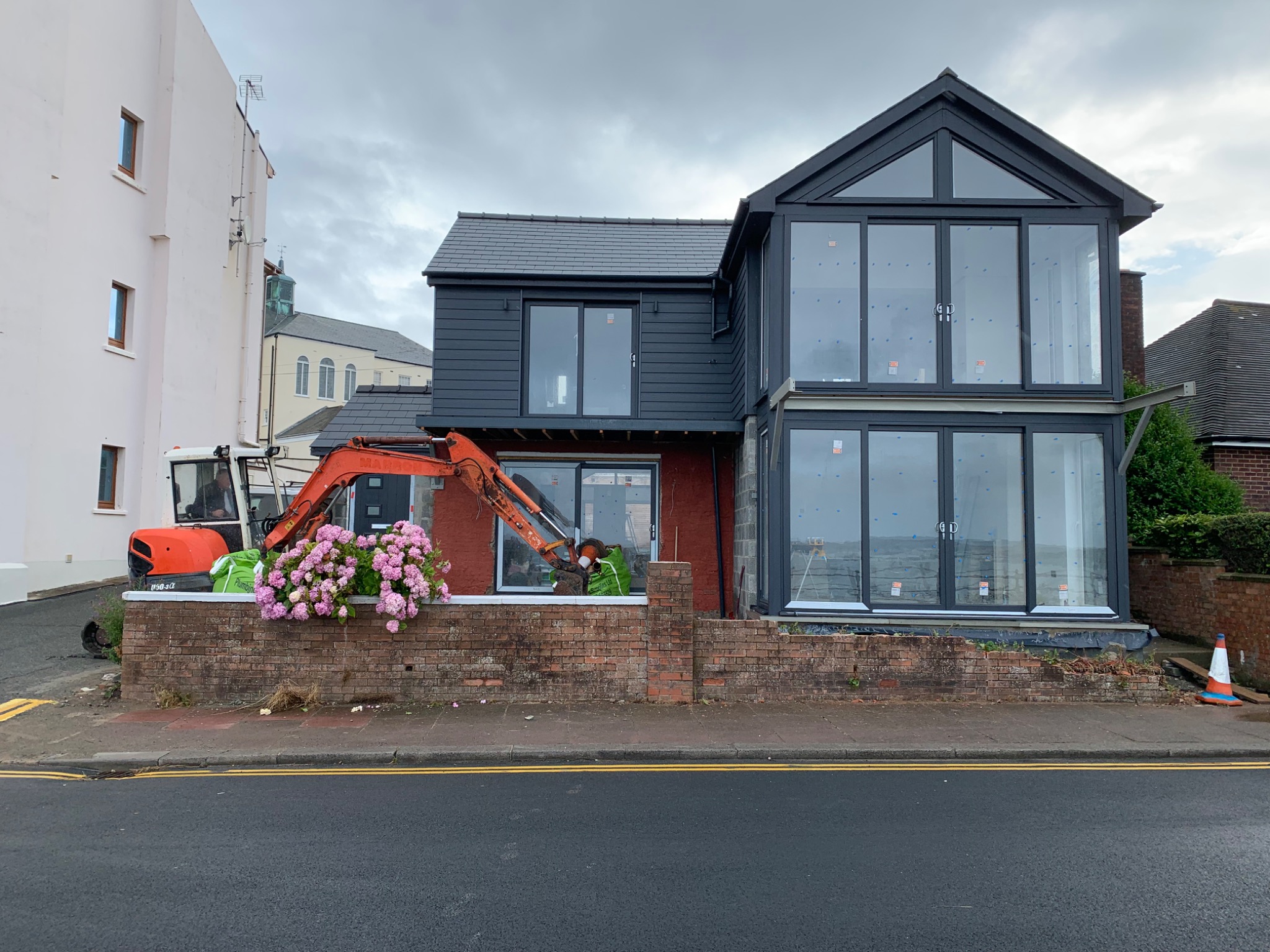 Dom w trakcie rozbudowy z użyciem małej koparki, widoczna murowana podmurówka i nowoczesna dobudówka z dużymi oknami.