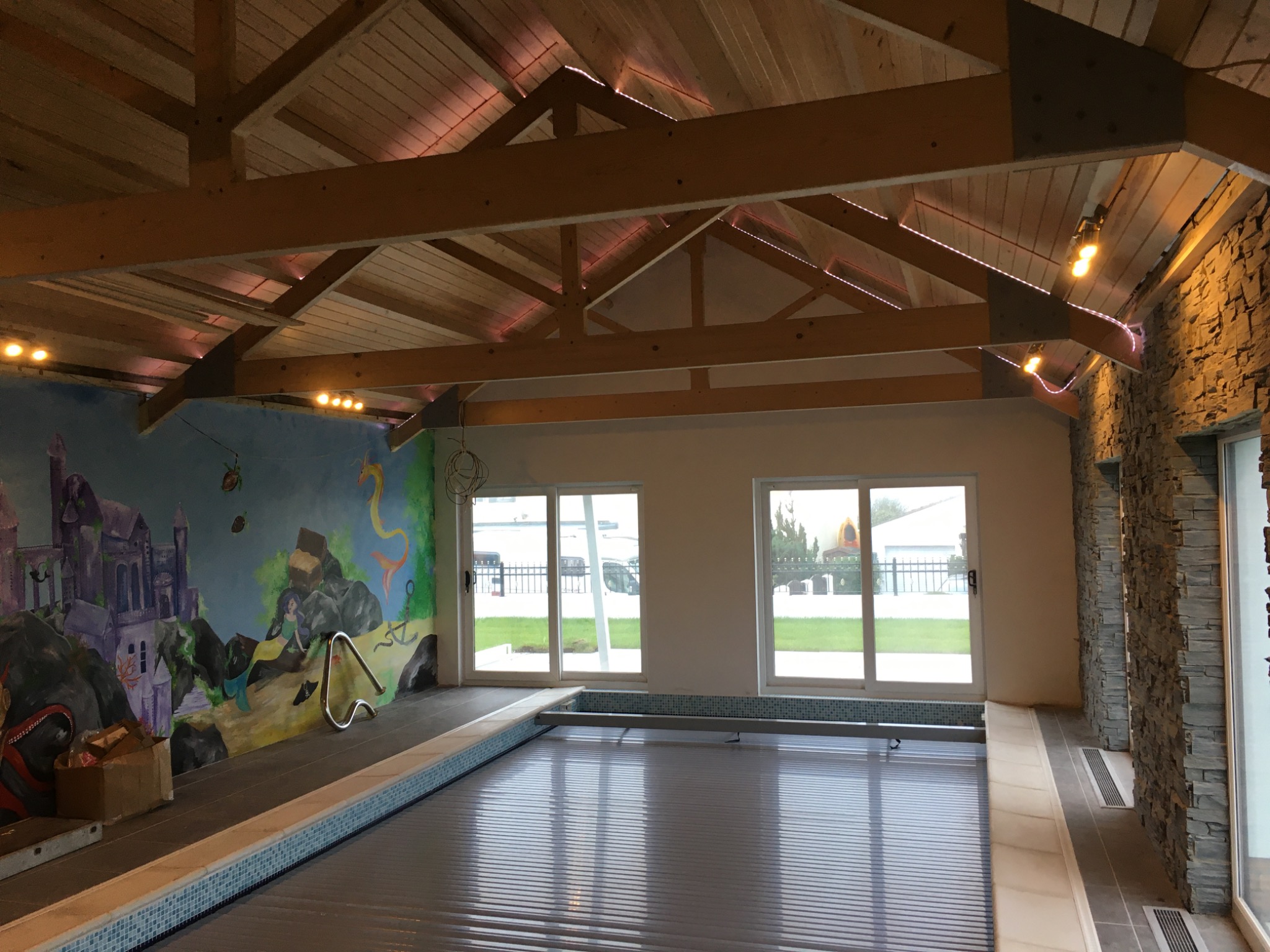 Wnętrze krytego basenu z drewnianą konstrukcją dachu, oświetleniem punktowym, muralem z motywami baśniowymi na ścianie oraz kamienną okładziną ściany bocznej.