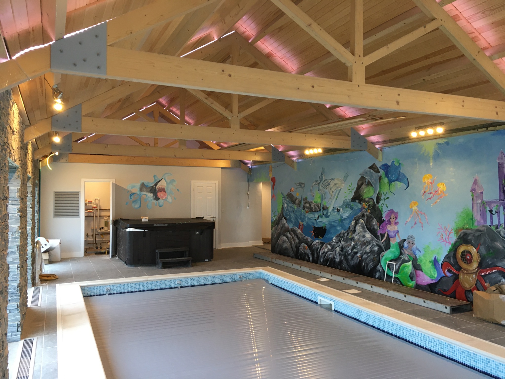Kryty basen z roletą, obok jacuzzi, ściana ozdobiona podwodnym muralem z syrenami i rekwizytami nurkowymi, drewniana konstrukcja dachu z oświetleniem.